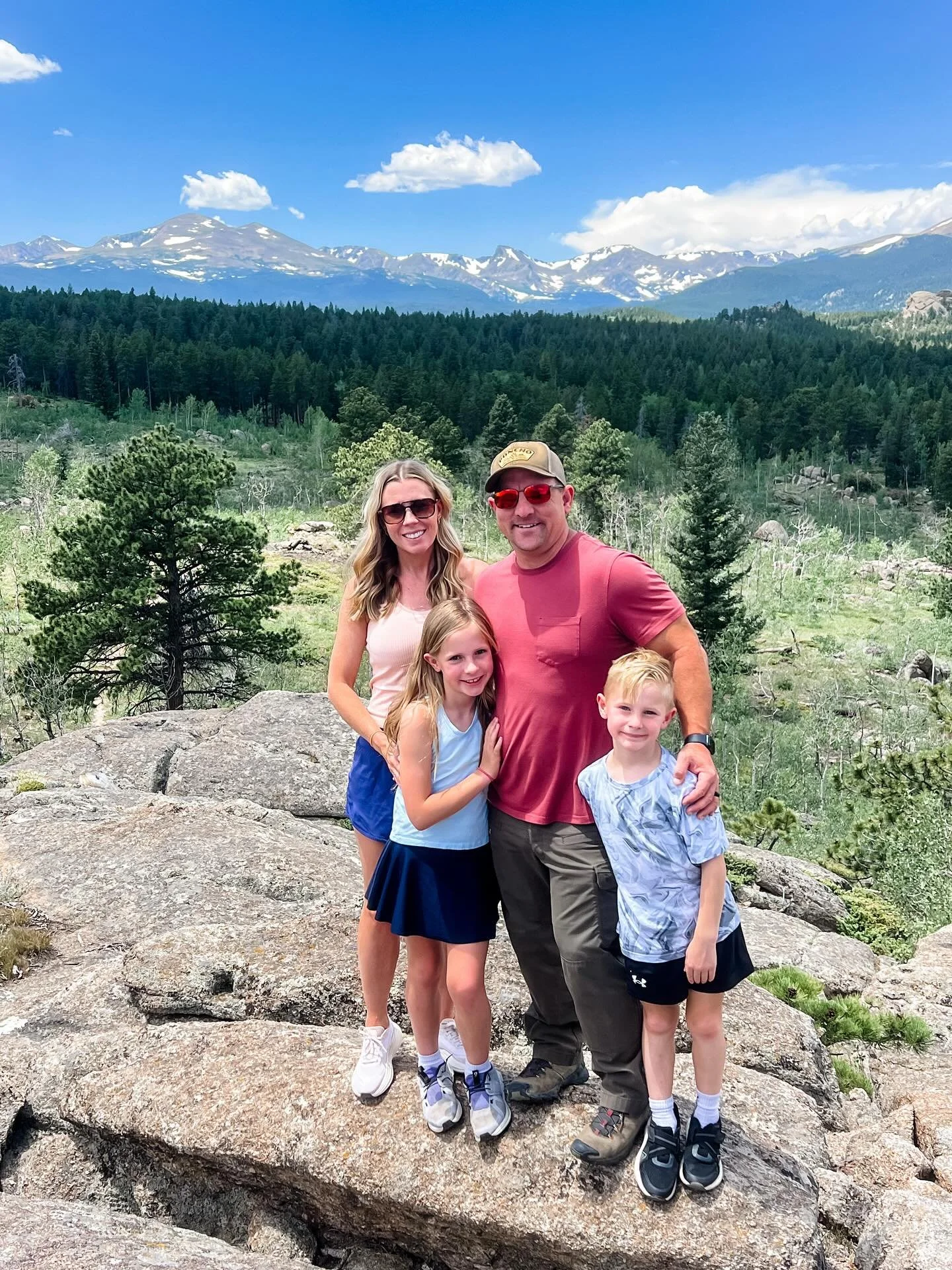 A family of four stands on a large rock with a backdrop of lush green trees and snow-capped mountains under a clear blue sky.