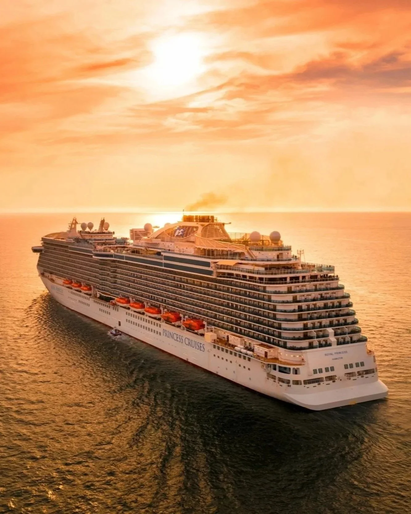 A large white cruise ship sailing on calm ocean waters during a sunset, with the sky glowing orange and pink, and the ship leaving a trail in the water.