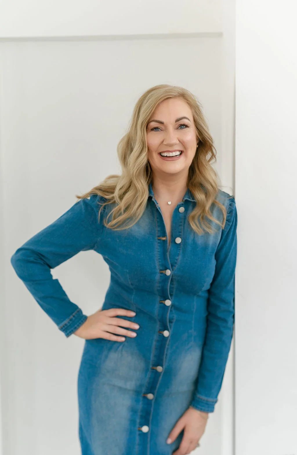 A woman with blonde wavy hair smiling, wearing a denim dress, standing against a white background.