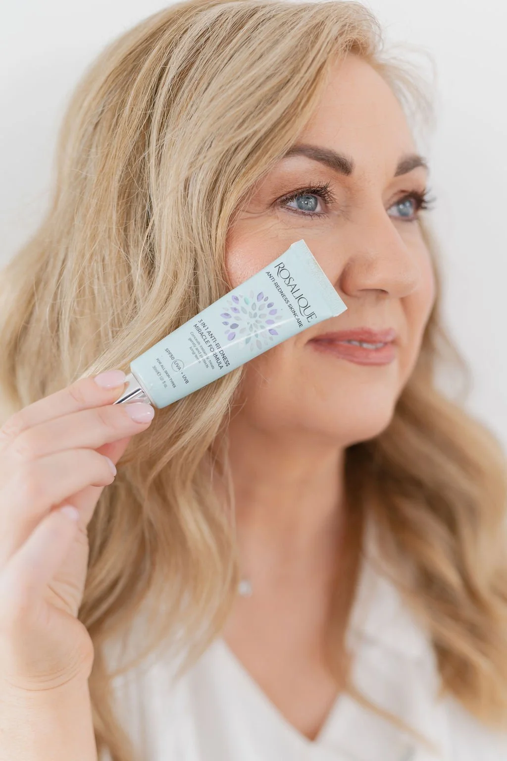 A woman with blonde hair and blue eyes applying skincare from a tube labeled "RosaliQue" to her face.