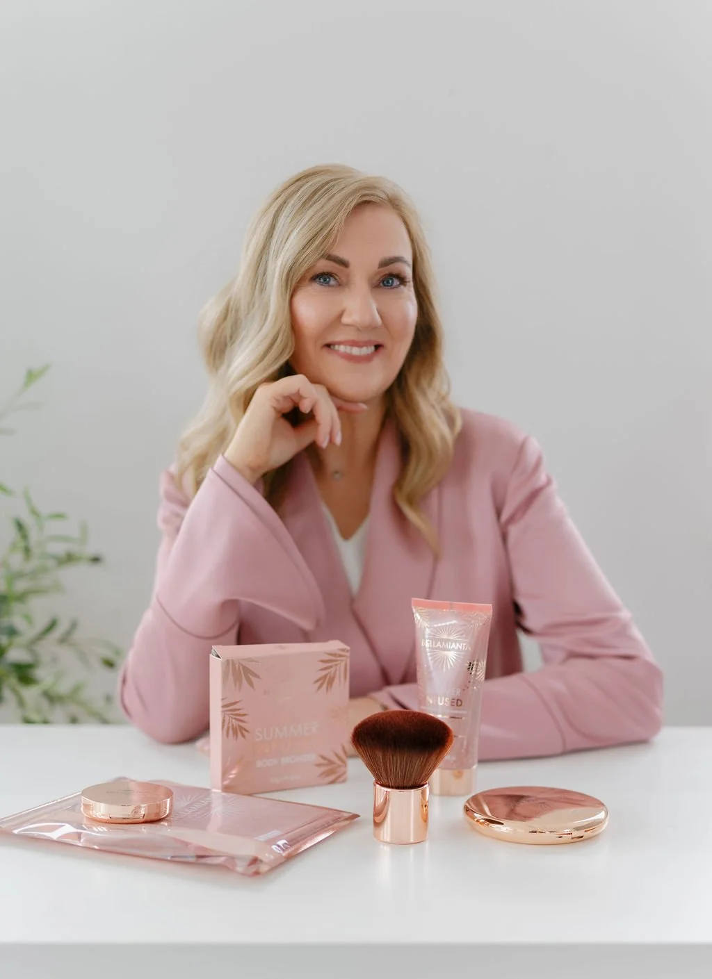 A woman with blonde wavy hair, smiling and resting her chin on her hand, sitting at a white table with pink and rose gold makeup products including a compact, brush, and tubes.