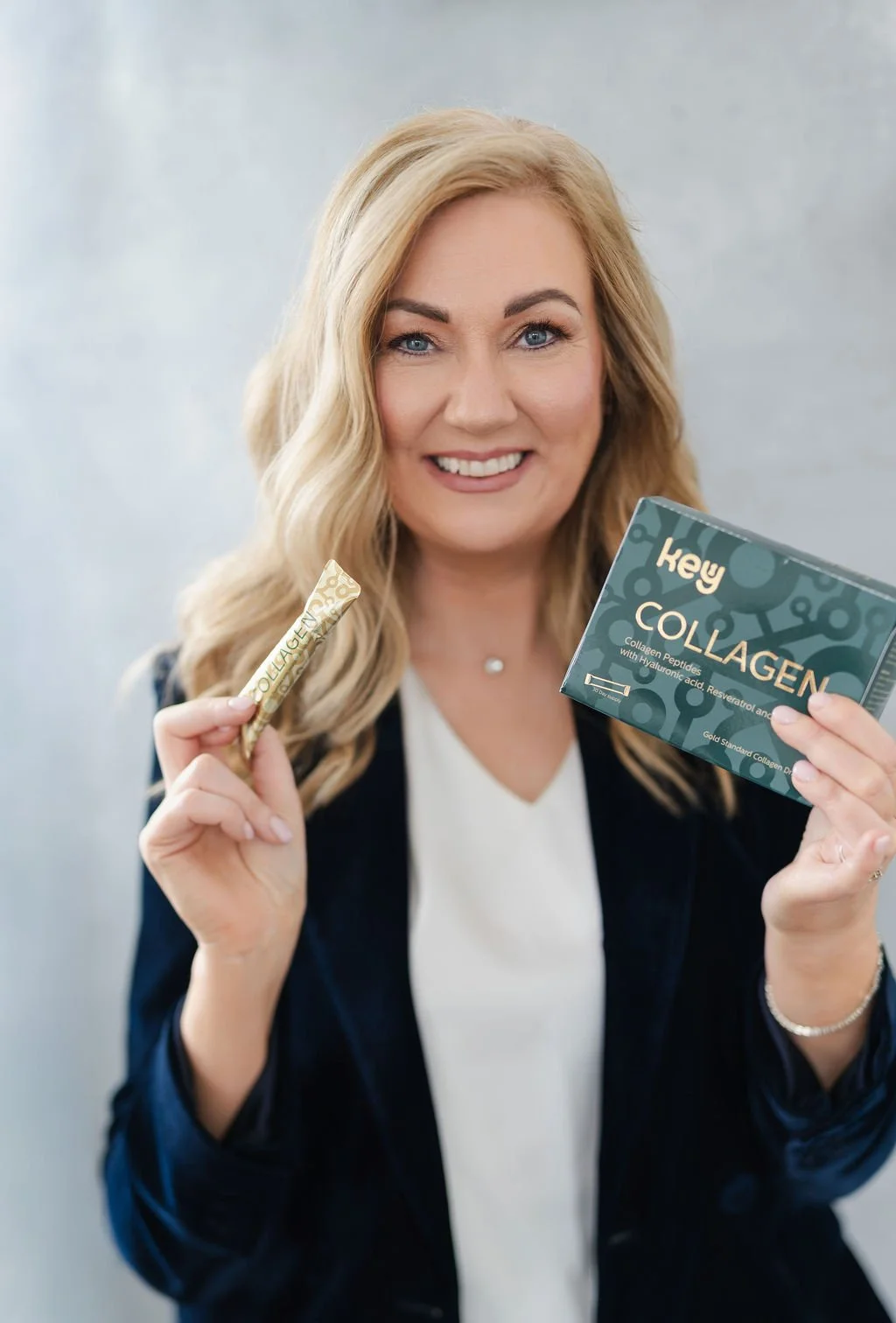 A woman with blonde hair smiling, holding a box of collagen supplement in her right hand and a single sachet of collagen in her left hand.