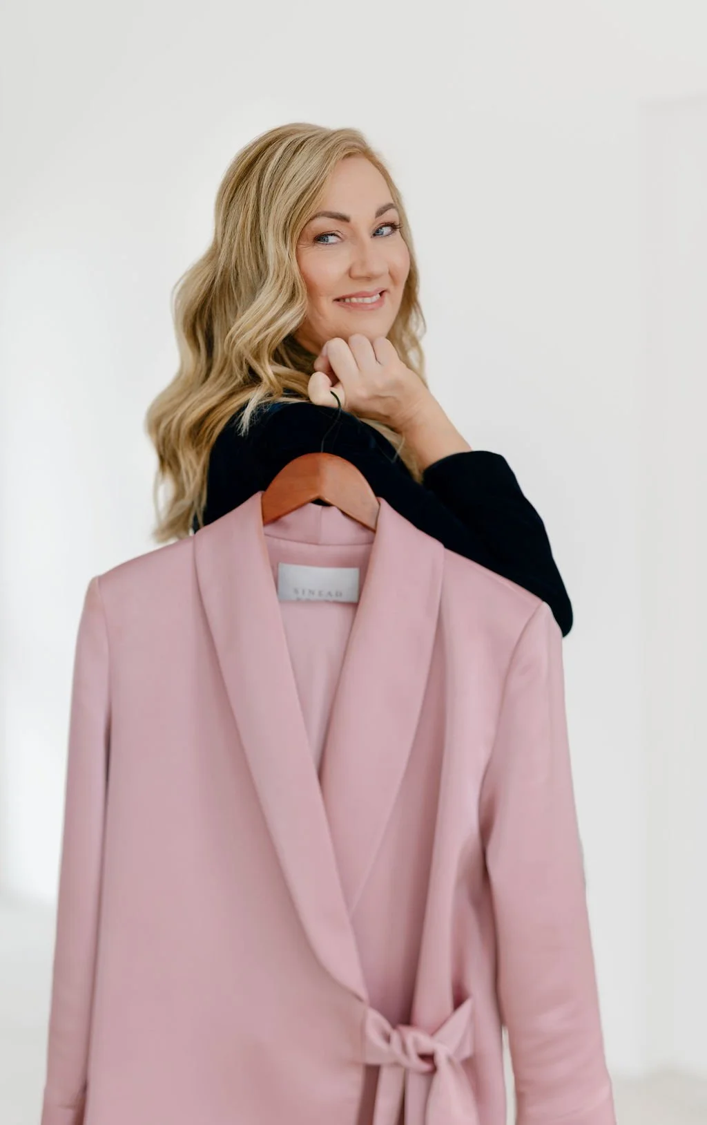 A woman with blonde hair and a black top holding a pink blazer on a wooden hanger against a white background.