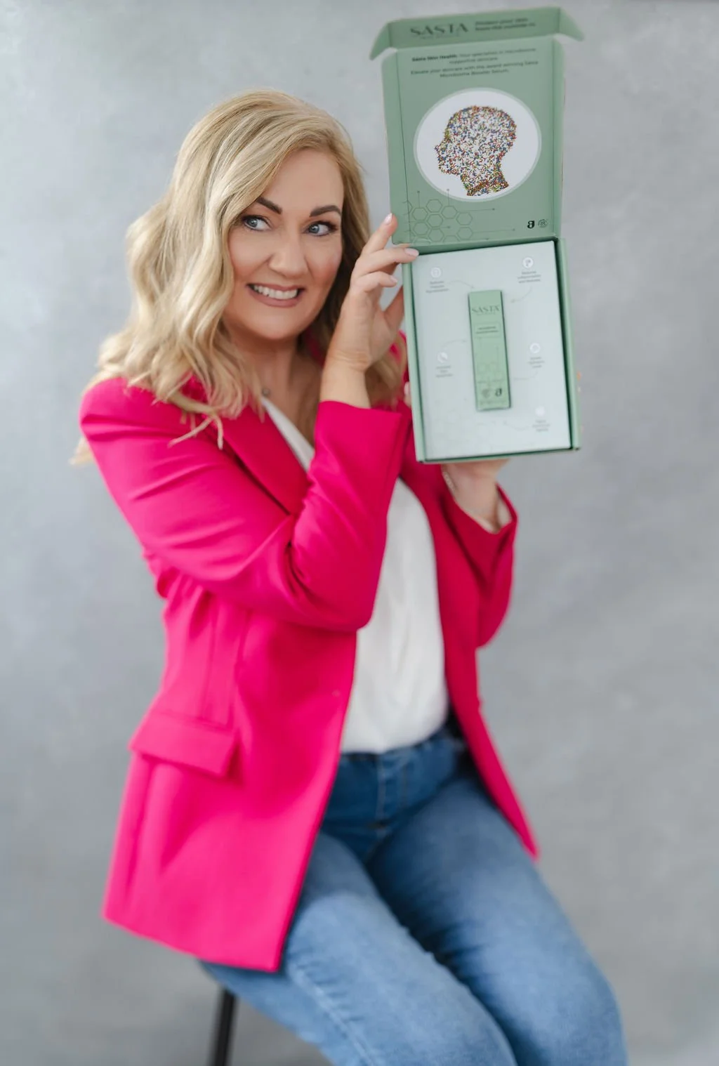 A woman with shoulder-length blonde hair in a pink blazer and white top sitting on a chair holding a green box with a product inside, smiling at the camera.