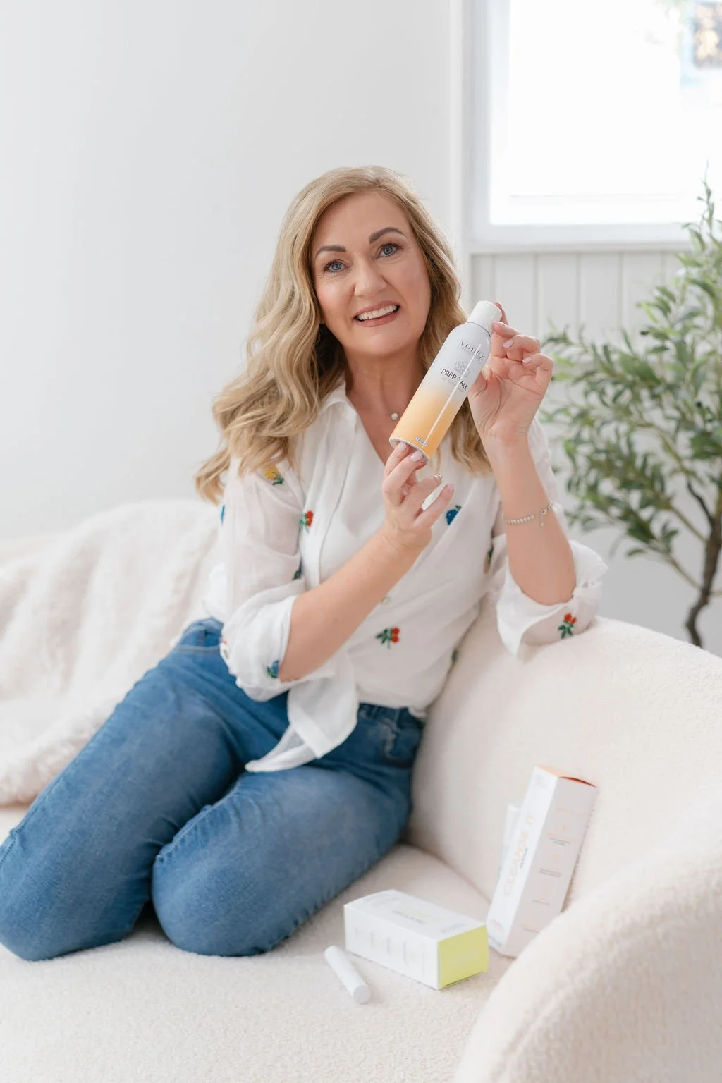 A woman with blonde hair and a white shirt sitting on a sofa in a bright room, holding a skincare product. There are skincare boxes and a tube on the sofa next to her.