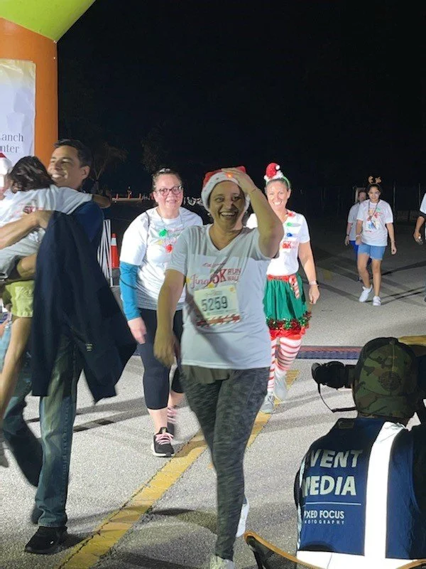 People participating in a nighttime charity race, wearing festive holiday attire including Christmas hats and costumes, with smiling faces and race bibs.