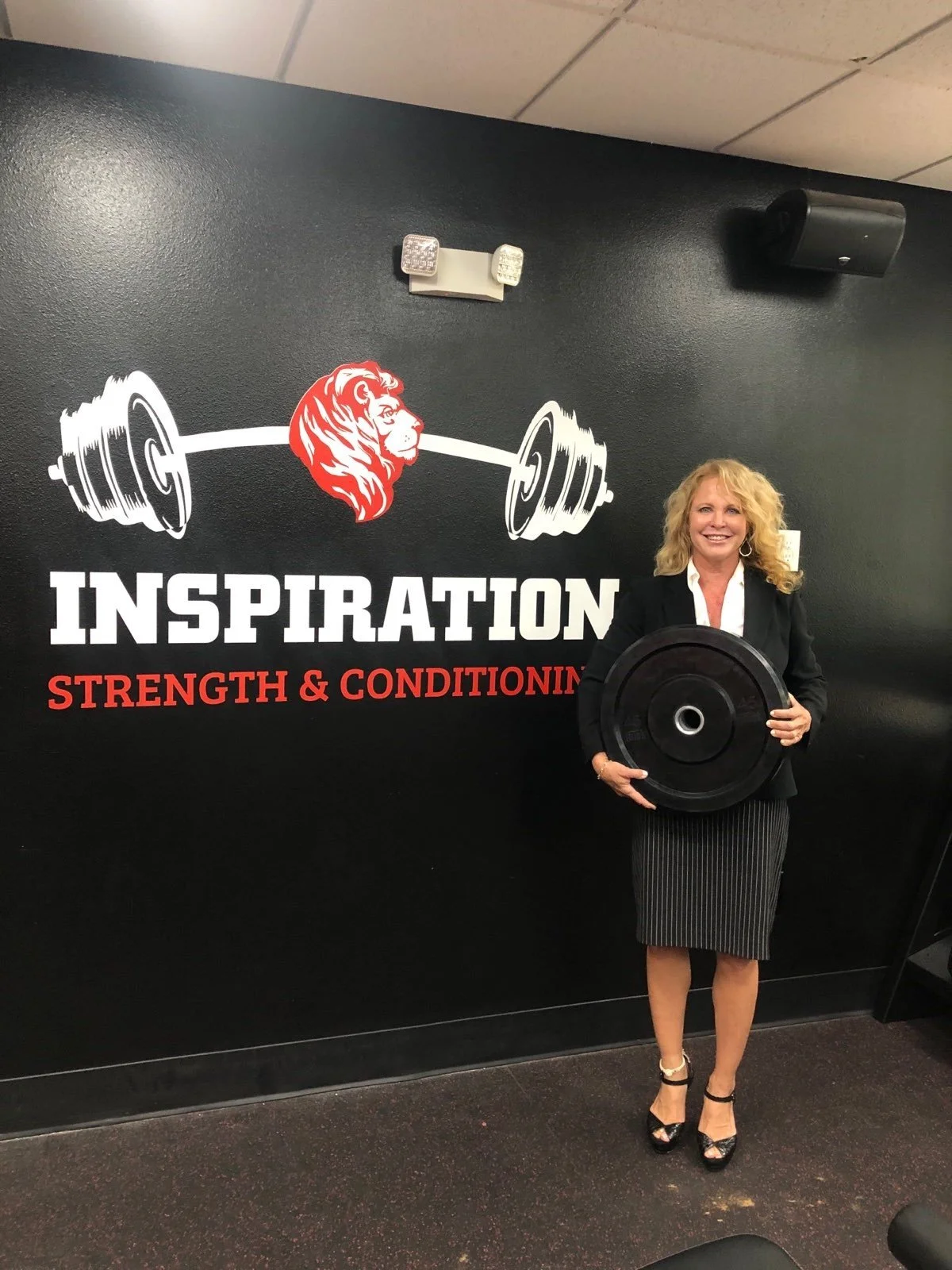 A woman in business attire holding a weight plate standing in front of a gym wall with the words "Inspiration Strength & Conditioning" and a lion logo.