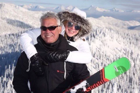 A man and a young girl in winter clothing hugging on a snowy mountain with snow-covered trees in the background, holding a snowboard.
