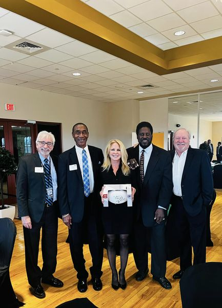 Five diverse professionals standing together in a reception or conference room, smiling, in formal business attire, with a woman in the center holding a white box.