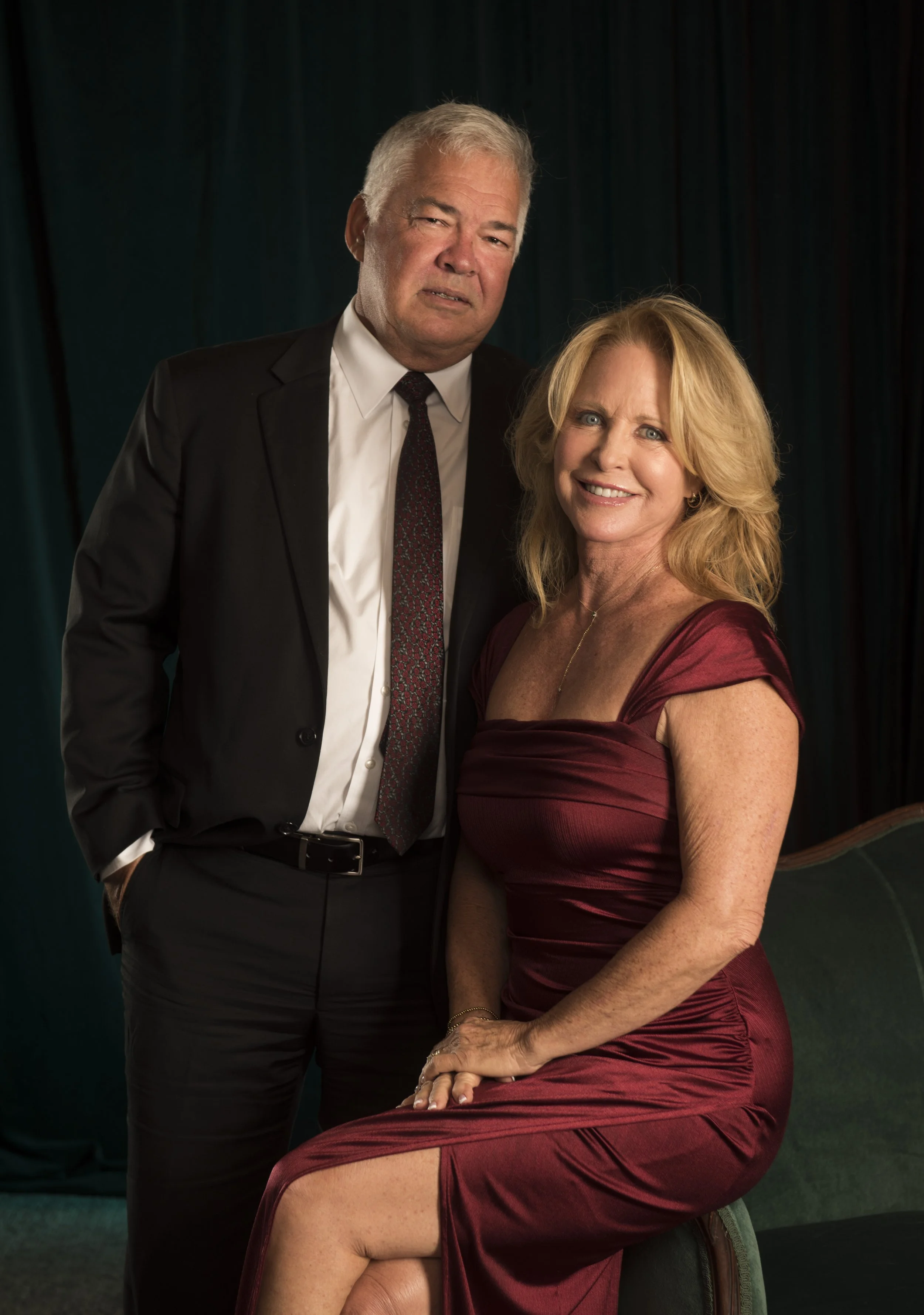 An older man with gray hair in a black suit and tie, and a smiling woman with blonde hair in a burgundy dress, posing together in front of a dark curtain background.