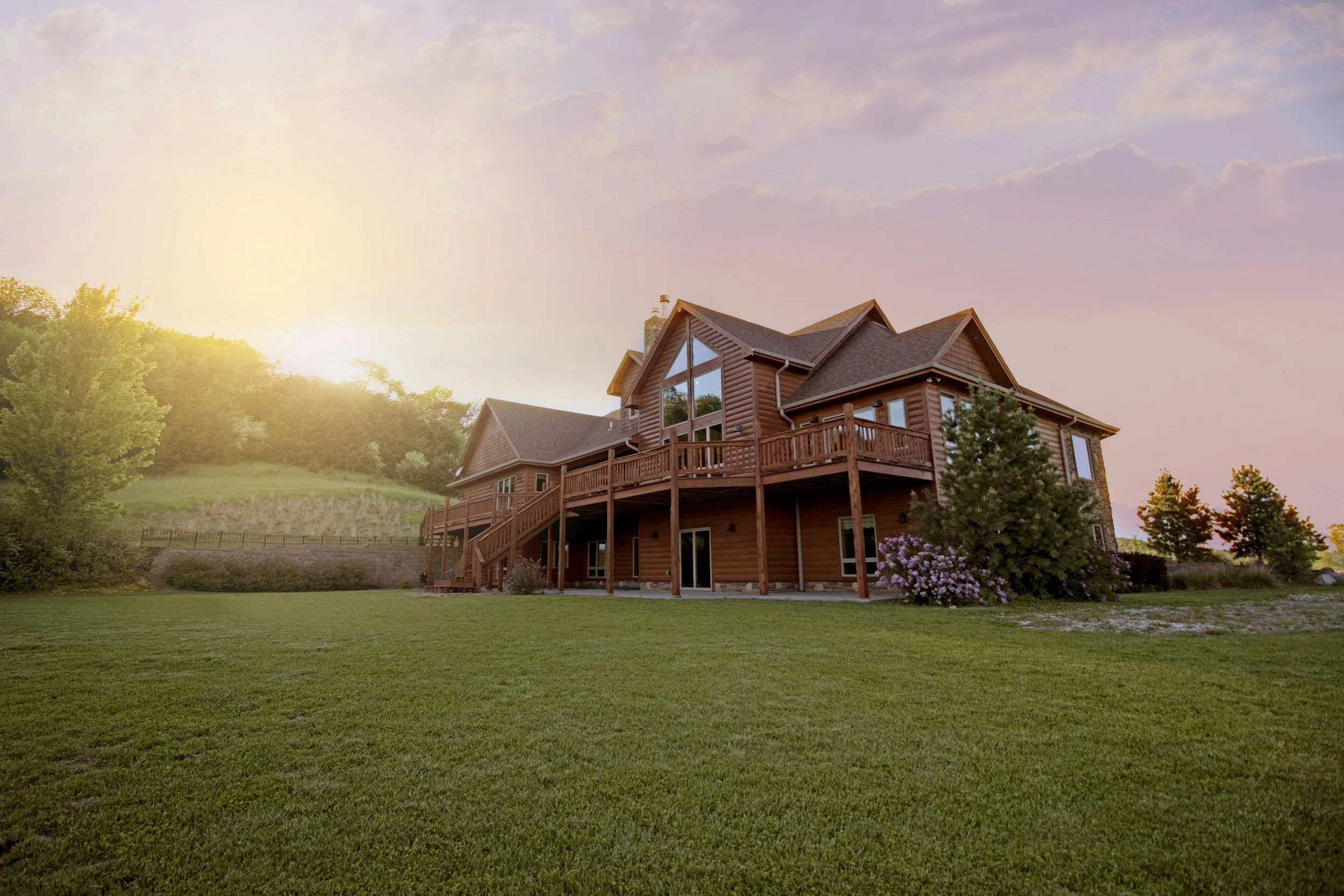 A large wooden house with a rooftop deck, surrounded by green grass, trees, and flowering bushes, at sunset.