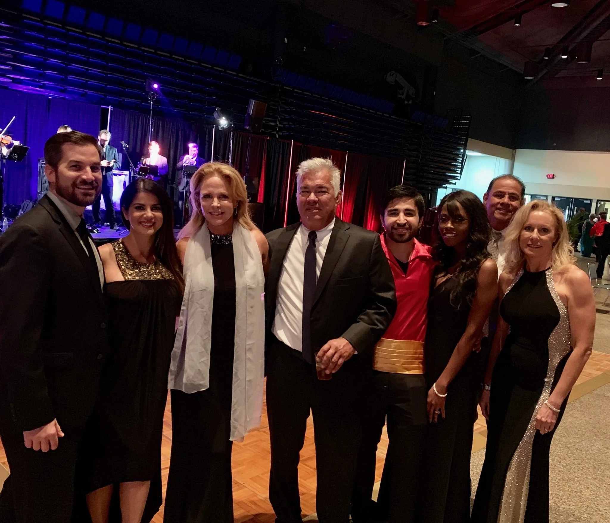 Group of eight people dressed in formal attire posing together at an indoor event or celebration with a live band performing on stage in the background.
