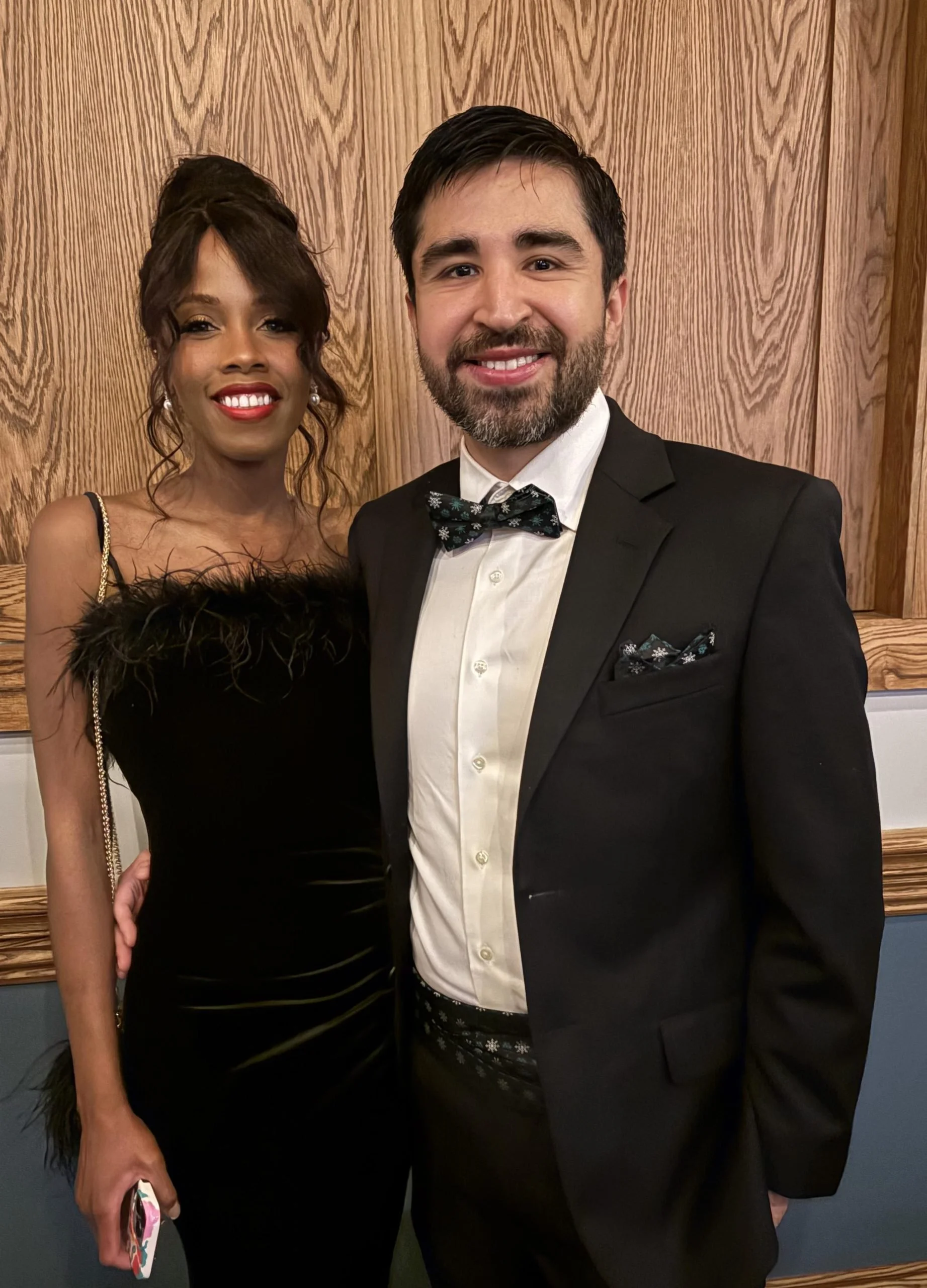 A man in a tuxedo smiling and standing next to a woman in a black dress, both posing for a photo in front of a wooden wall.