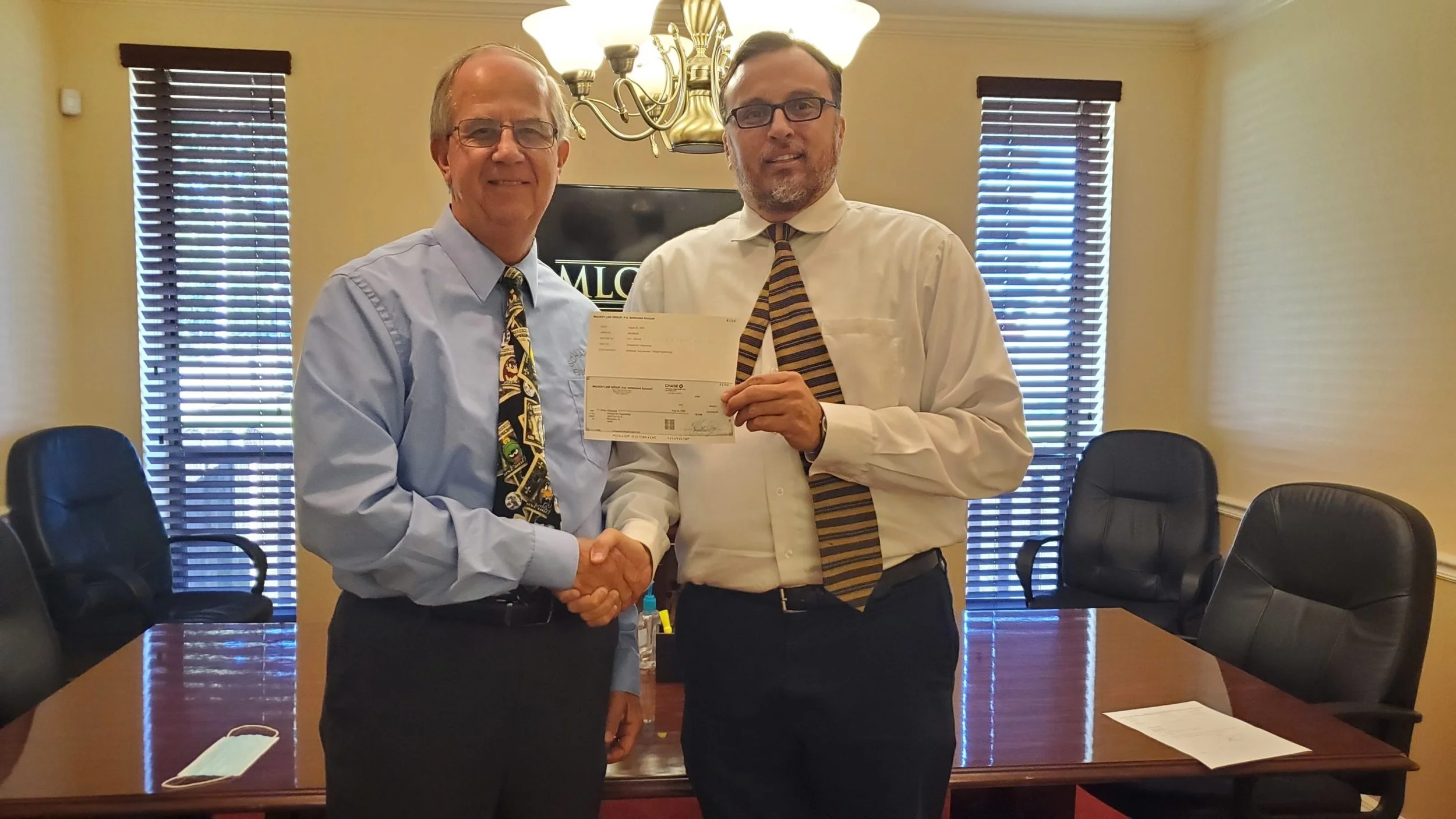 Two men in dress shirts and ties shaking hands in a conference room, with one holding a check. There are black chairs and a table with papers and a face mask on it, and windows with blinds behind them.