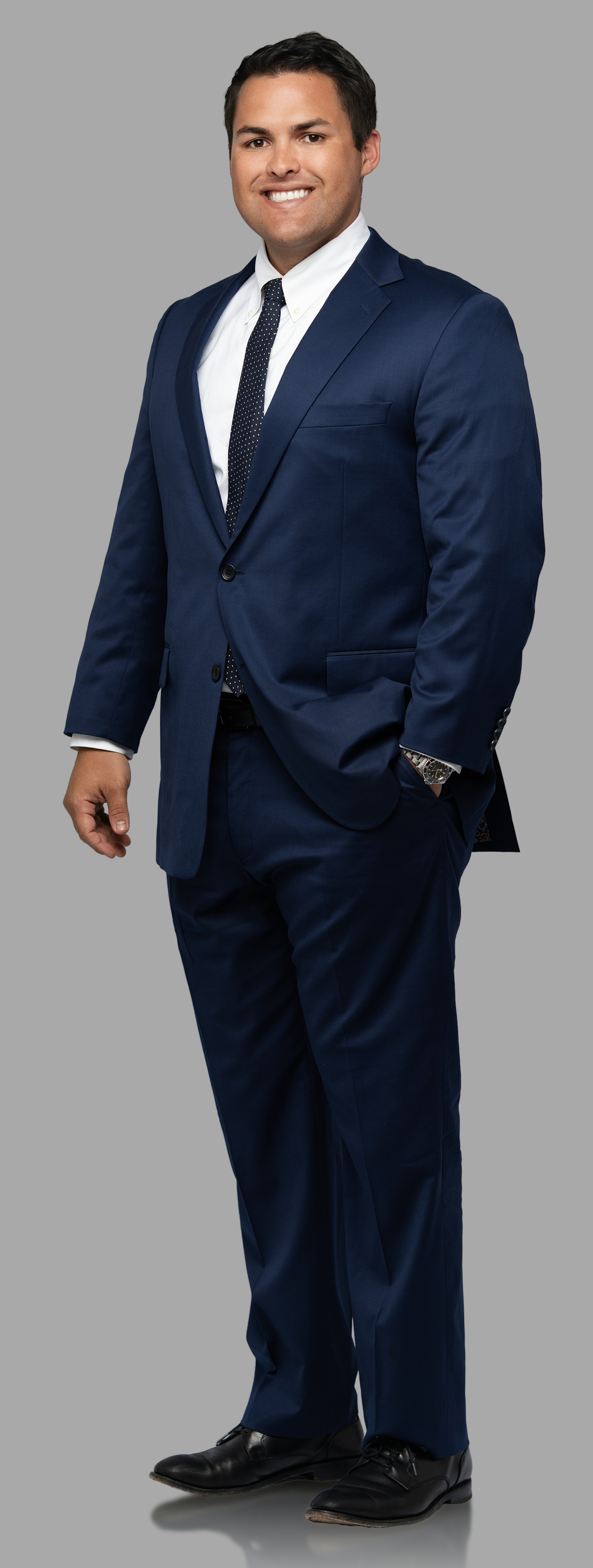 A man in a navy blue suit with a white shirt and a dotted black tie, standing against a gray background, smiling with one hand in his pocket and looking at the camera.