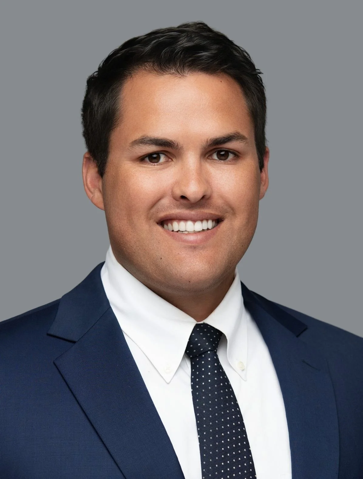 A professional headshot of a young man with dark hair, smiling, wearing a navy suit, white shirt, and a navy tie with white dots against a gray background.