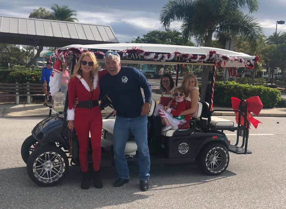 People celebrating Christmas standing next to a decorated golf cart with festive decorations and a large red bow, outdoors with palm trees and overcast sky.