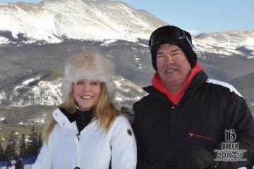 A smiling woman and man in winter clothing outdoors with snow-covered mountains in the background.