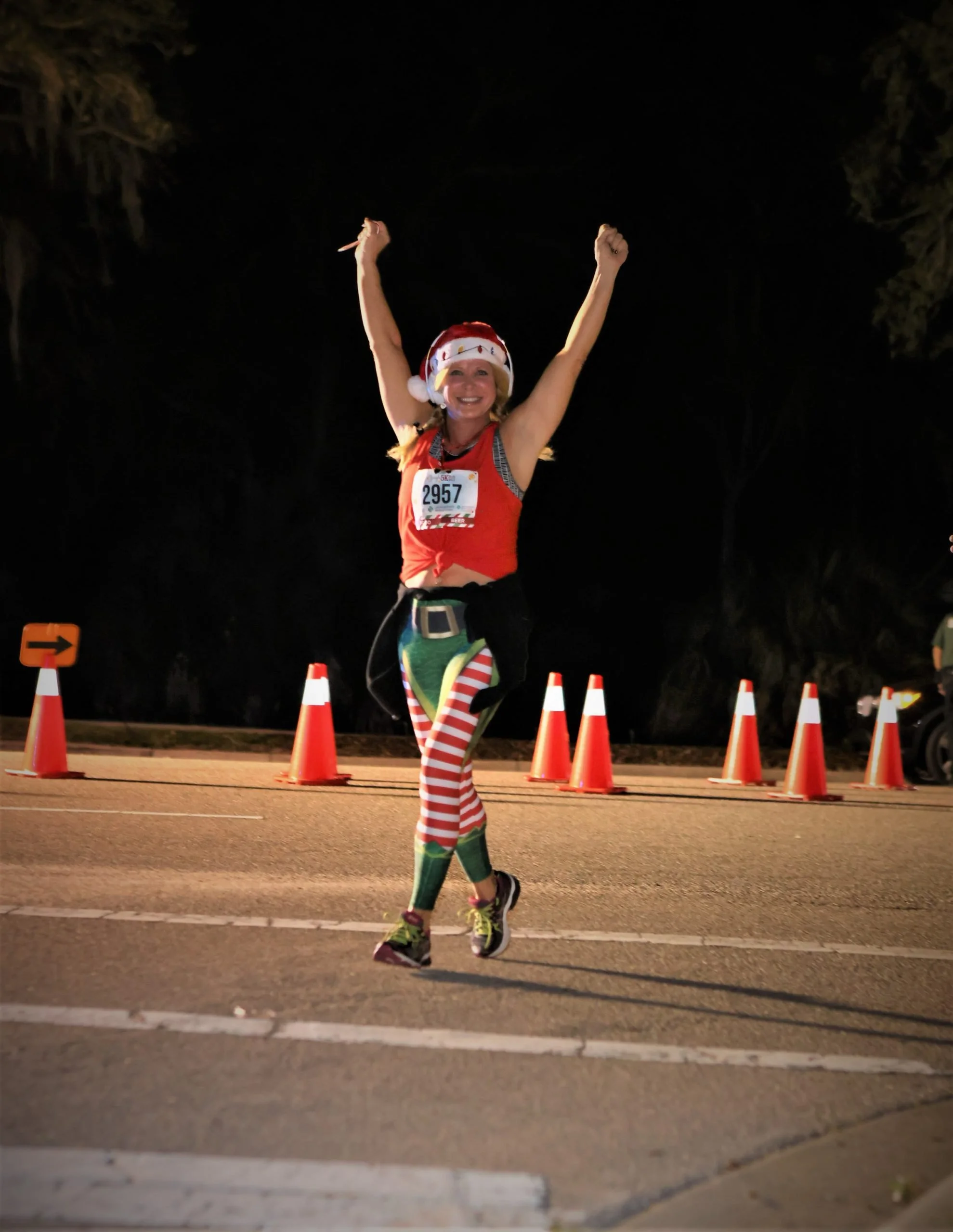 A woman in festive clothing and a Santa hat crossing a finish line during a nighttime race, cheering with her arms raised.