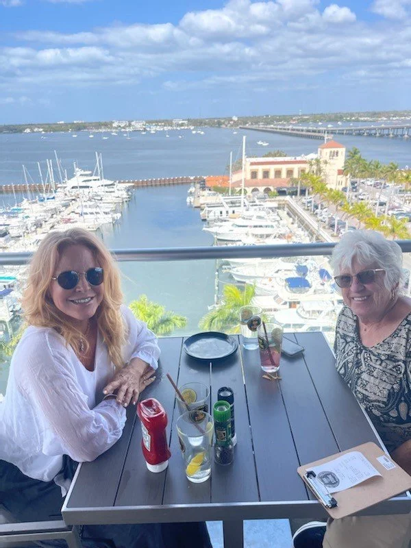 Two women sitting at a table with drinks, overlooking a marina with boats and a body of water with a sky filled with clouds.