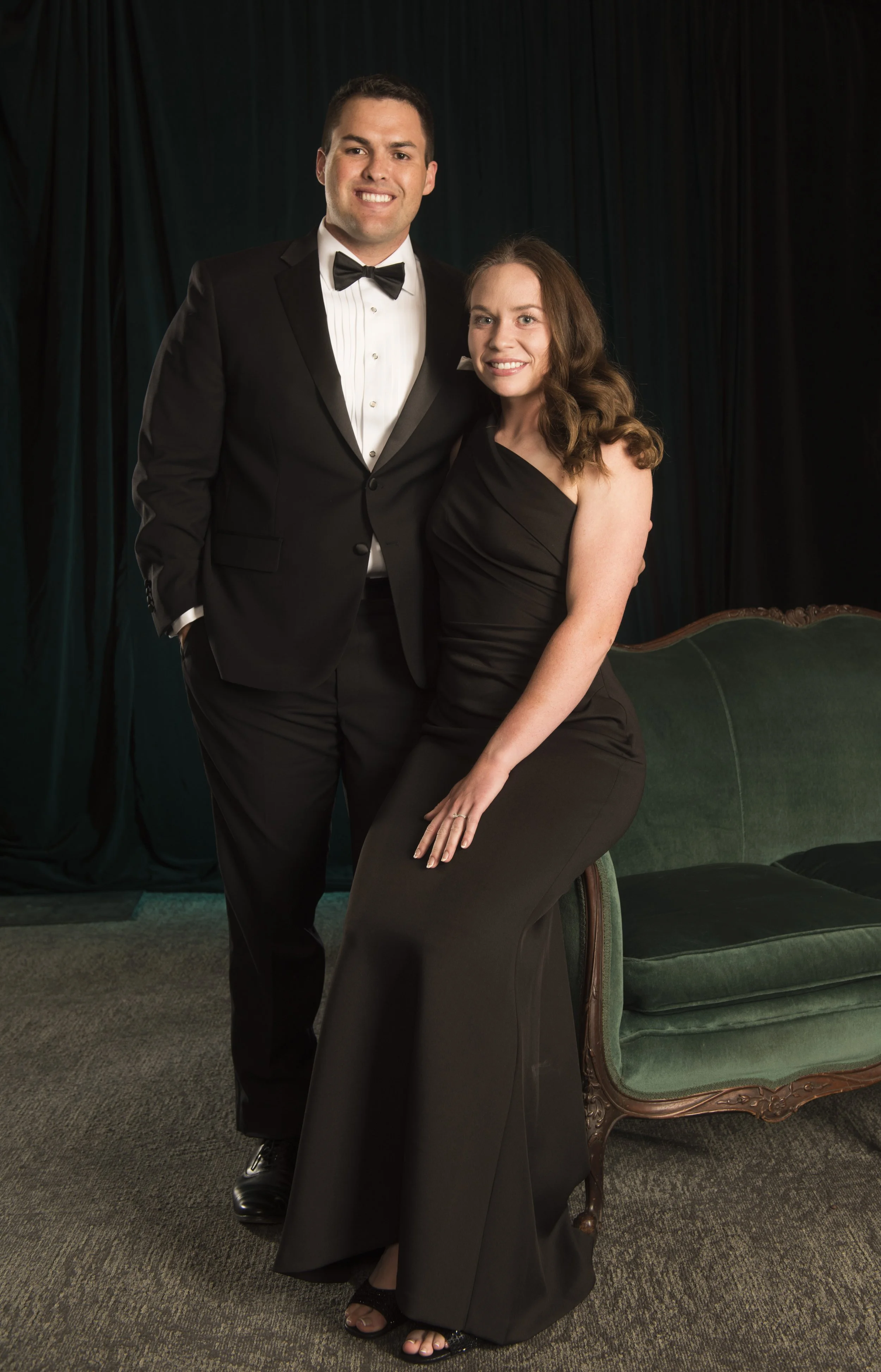 A man dressed in a tuxedo and bow tie standing next to a woman in a black evening gown, sitting on a green velvet vintage-style couch in front of a dark curtain backdrop.