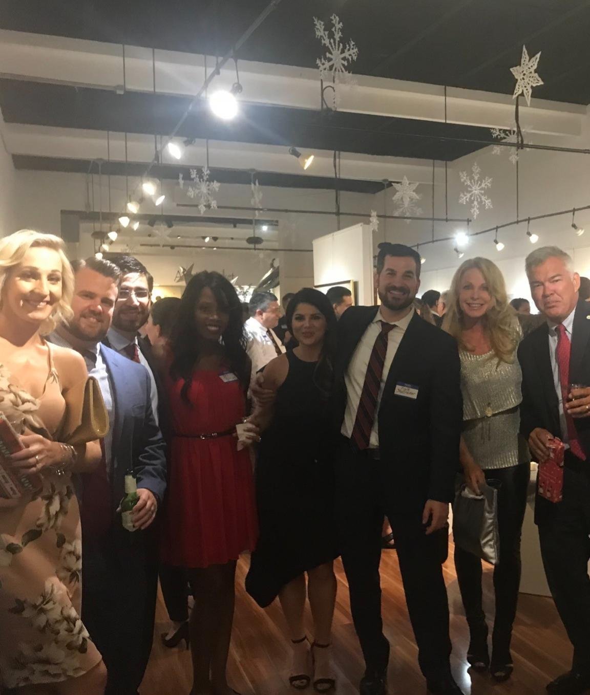 A diverse group of people dressed in formal attire at a holiday celebration, with Christmas and winter decorations hanging from the ceiling.