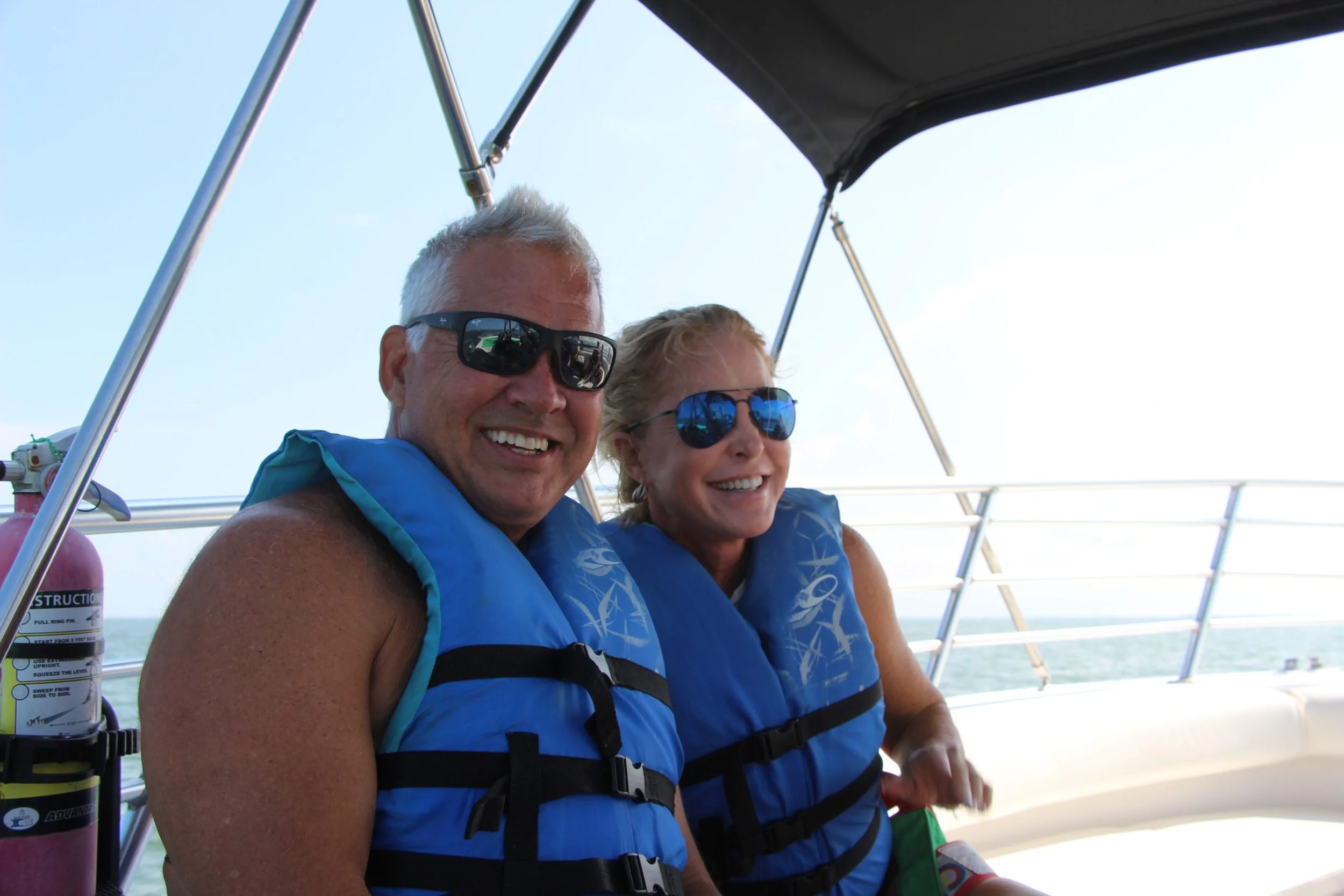 Two people, a man and a woman, sitting close together on a boat, both smiling and wearing sunglasses and life jackets.