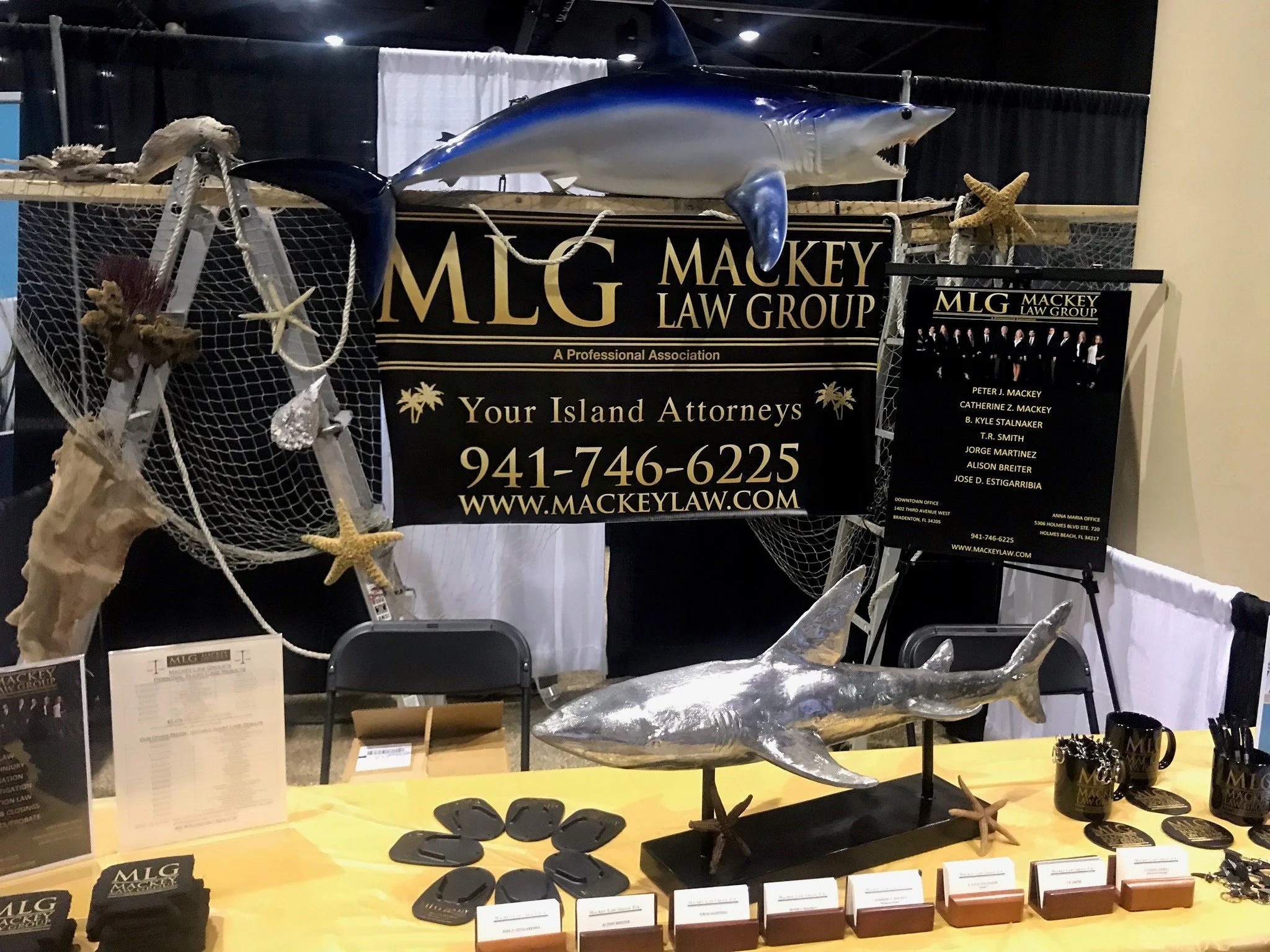 Display booth with a large mounted model of a shark, conference banners for Mackey Law Group, decorative sea starfish, shells, and fishing-themed items.