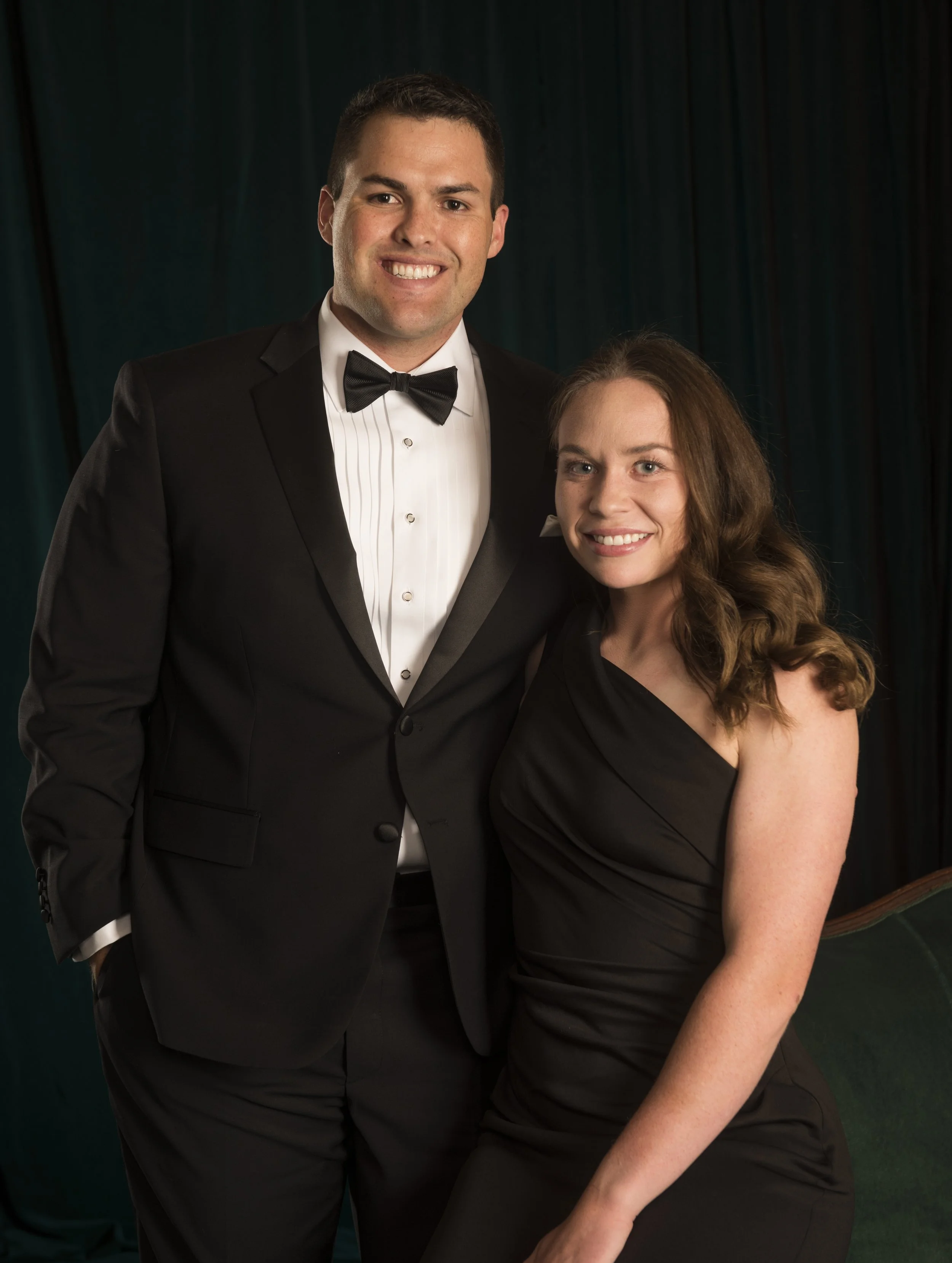 A man in a tuxedo and a woman in a black dress smiling together at a formal event or celebration.