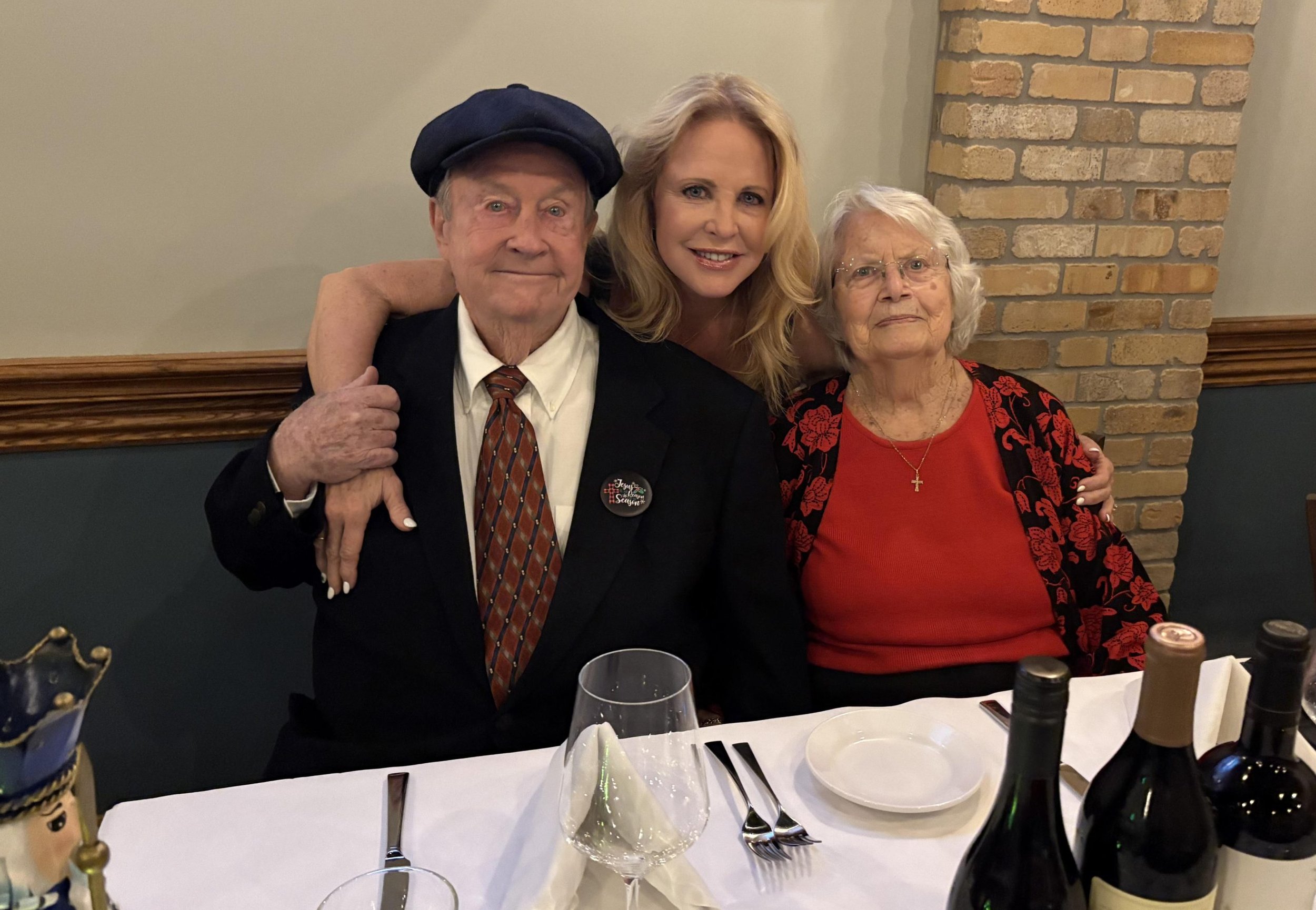 Three people sitting at a dining table, including an elderly man with a black hat and a button reading 'Joyful Celebrations', a woman with blonde hair, and an elderly woman with glasses and a red top. The table has wine glasses, bottles of wine, and 