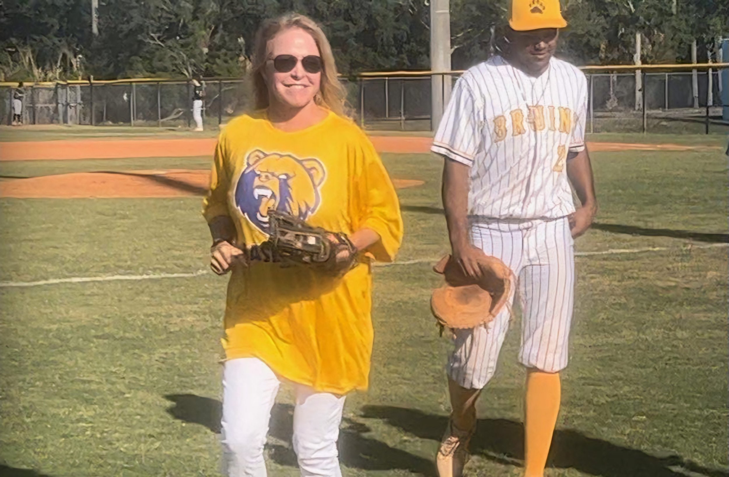 A woman in a yellow sports jersey and sunglasses smiling while walking on a baseball field, next to a man in a baseball uniform and helmet, on a sunny day.