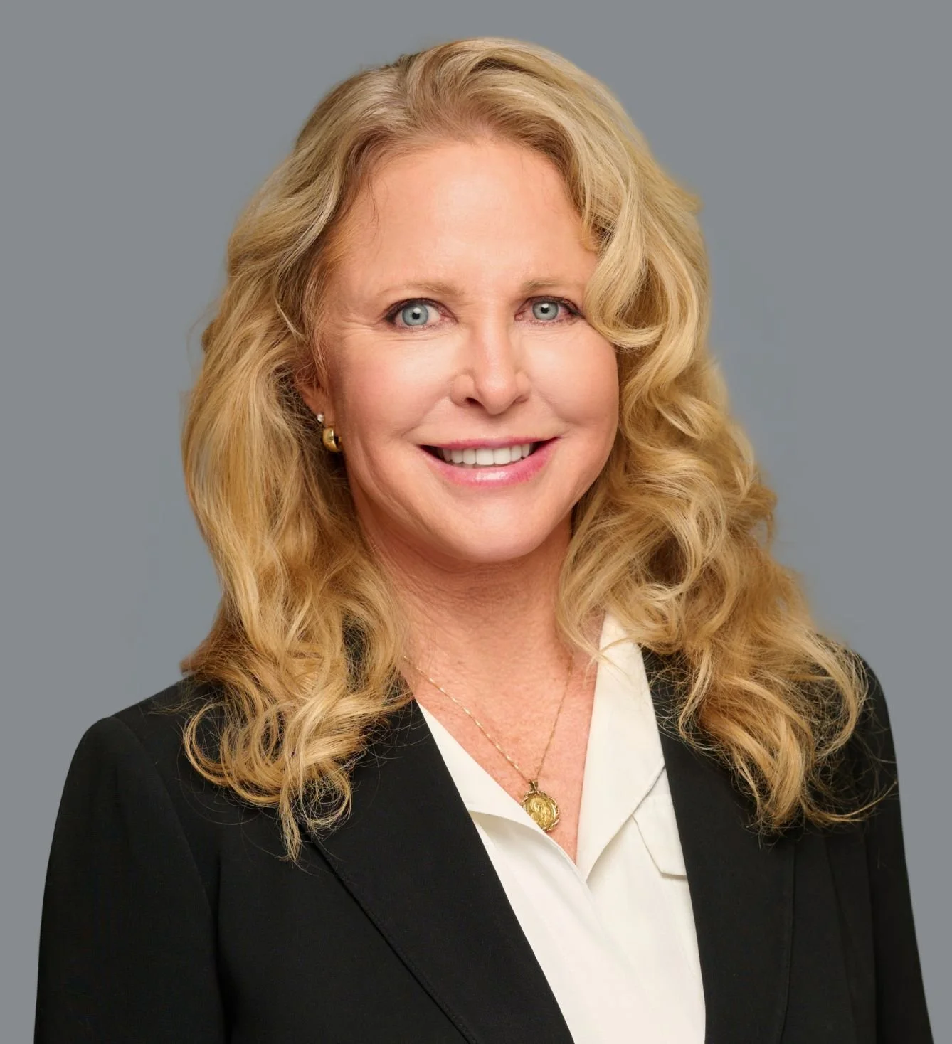 A professional woman with long, curly blonde hair, blue eyes, and a warm smile, dressed in a black blazer and white blouse, wearing gold jewelry, against a plain gray background.