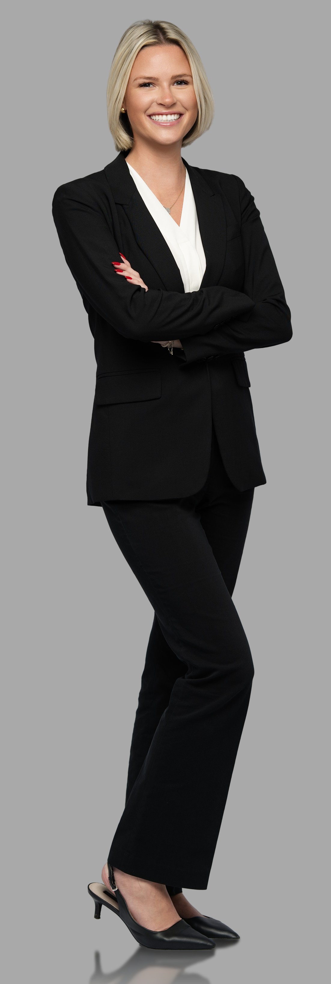 A professional woman in a black business suit with a white blouse, posing with arms crossed and smiling. She has blonde hair, red nail polish, and is wearing black high-heeled shoes.