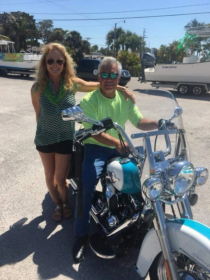 A woman with blonde hair, sunglasses, and a black and white chevron top, stands next to a man sitting on a blue and white motorcycle. The man has gray hair, wears sunglasses, a neon green shirt, and blue jeans. They are outdoors, with boats and trees