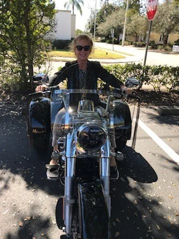 Woman with blonde hair wearing sunglasses, sitting on a black motorcycle in a parking lot, smiling at the camera.