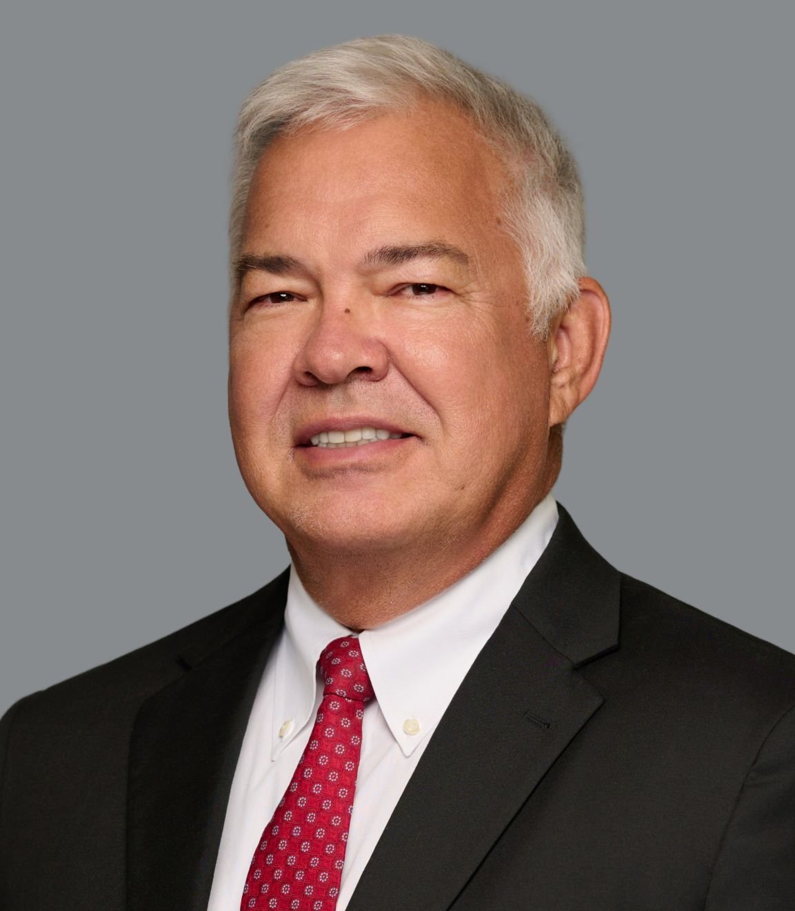 A middle-aged man with gray hair, wearing a black suit, white shirt, and red tie, smiling against a gray background.