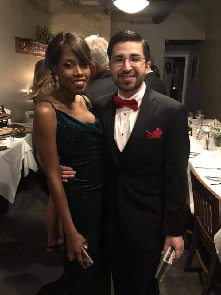 A woman in a dark green evening gown and earrings stands next to a man in a black tuxedo with a red bow tie and pocket square. They are smiling and posing for a photo at a formal event, with tables set with white tablecloths, plates, glassware, and f