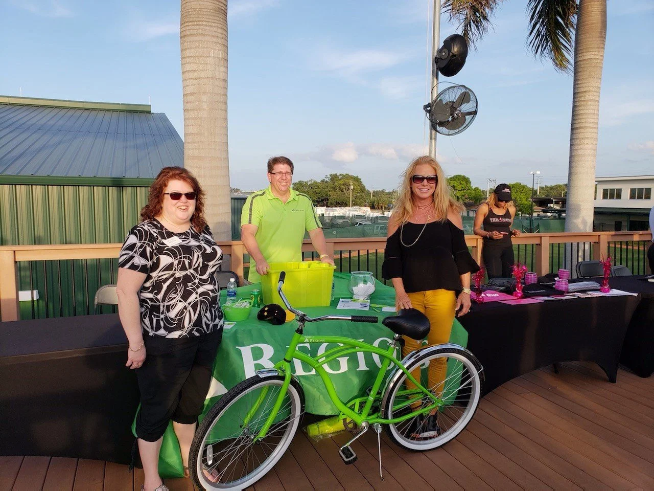 Three women and one man smiling at an outdoor event, with a green bicycle, tables with pink decorations, and palm trees in the background.