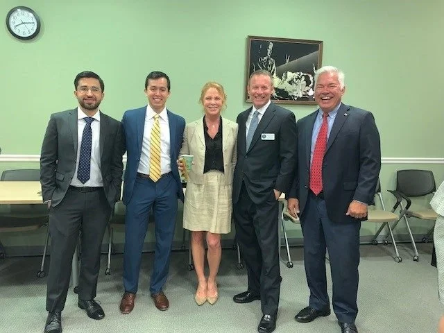 Five professionals standing in a meeting room, posing for a photo. The group includes three men in suits and a woman in a beige skirt and blazer, all smiling. There is a clock on the wall and a framed black-and-white photo behind them.