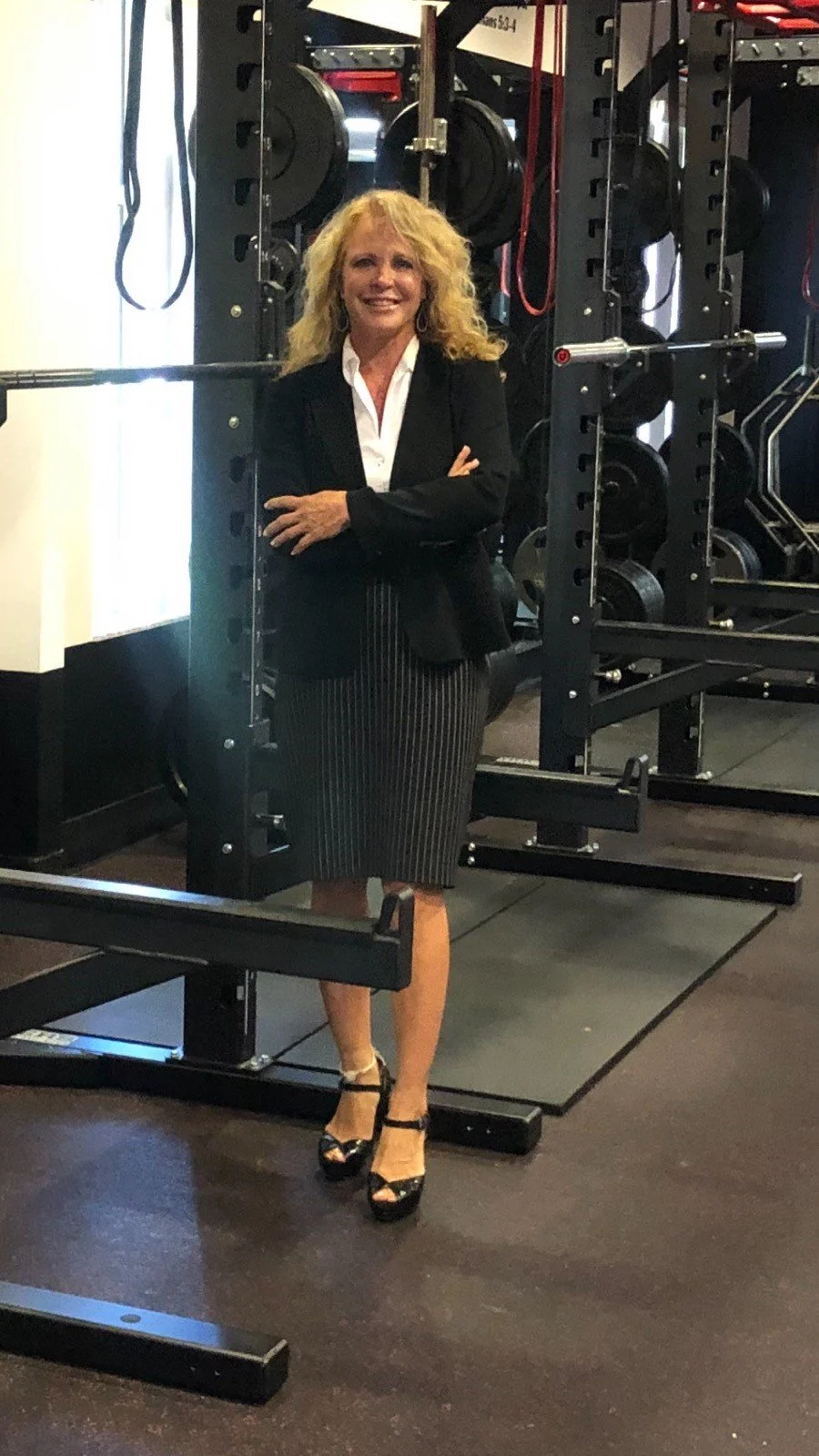 A woman standing in a gym with weights and workout equipment in the background, dressed in a black blazer, white shirt, striped skirt, and black high heels.