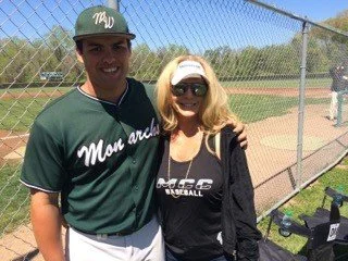A young man in a green baseball uniform and a woman in sportswear standing together at a baseball field, with a chain-link fence and other people in the background.