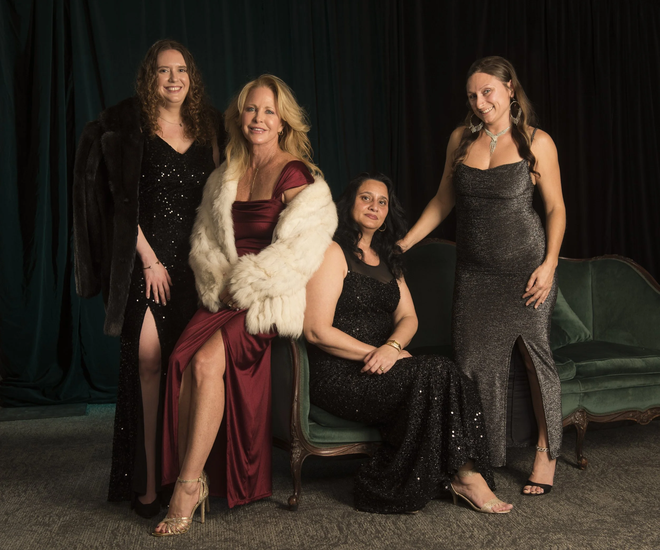 Group of five women in glamorous evening attire, some sitting and some standing, in front of dark curtains, posing for a photo at a formal event.