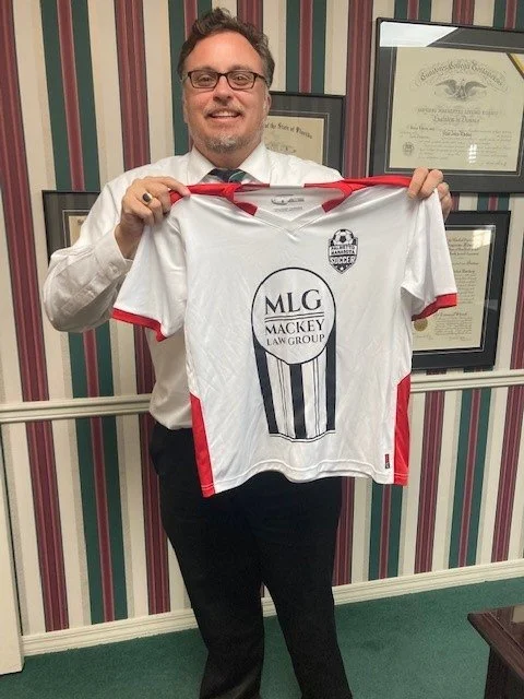 A man with glasses and a beard, dressed in a white shirt and black pants, holding a soccer jersey with red accents. The jersey features a logo with the text 'MLG Mackey Law Group' and a soccer emblem. The background includes framed documents on strip
