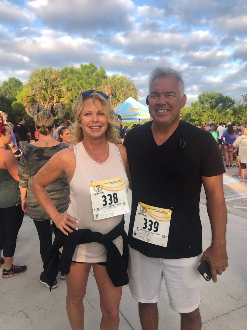 Two people with race bibs smiling at a race event, woman in tank top and shorts, man in black shirt and white shorts, crowd in background, trees, partly cloudy sky.