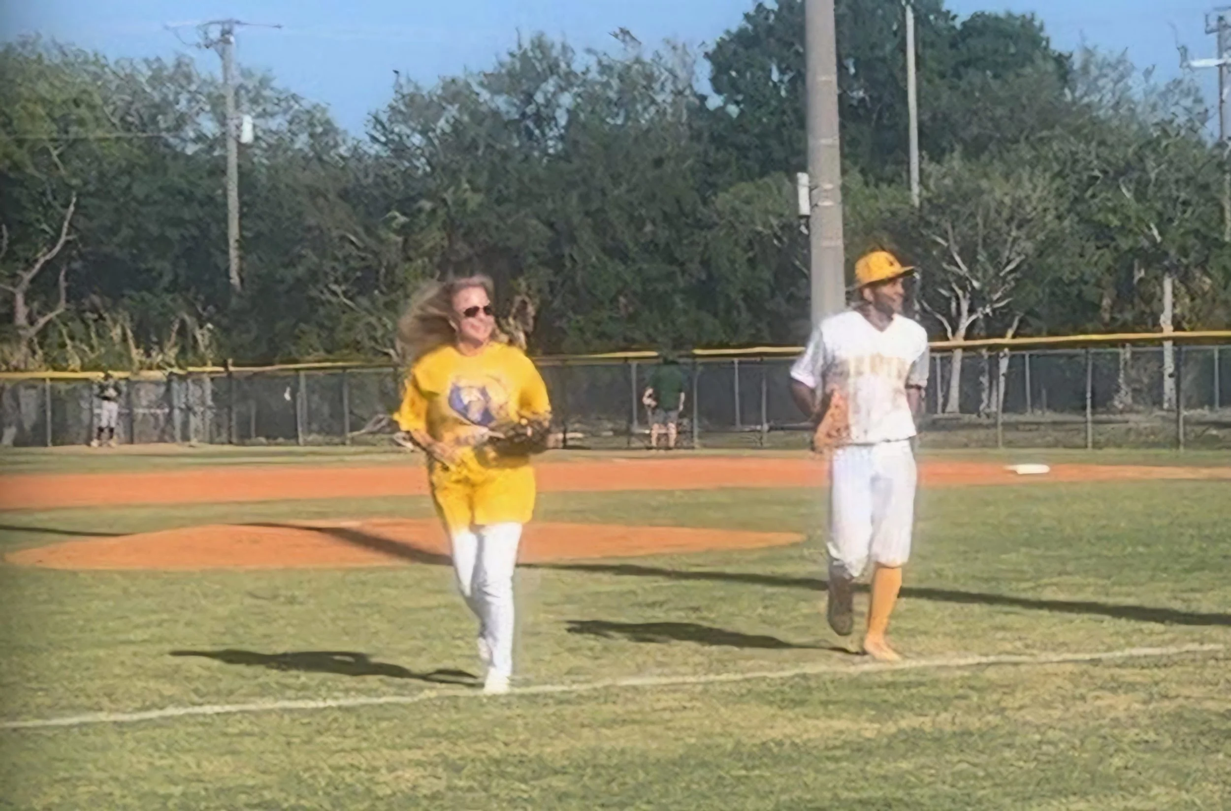 Two women on a baseball field, one in a yellow sports jersey and the other in a white uniform, walking or running near the base with a fence and trees in the background.