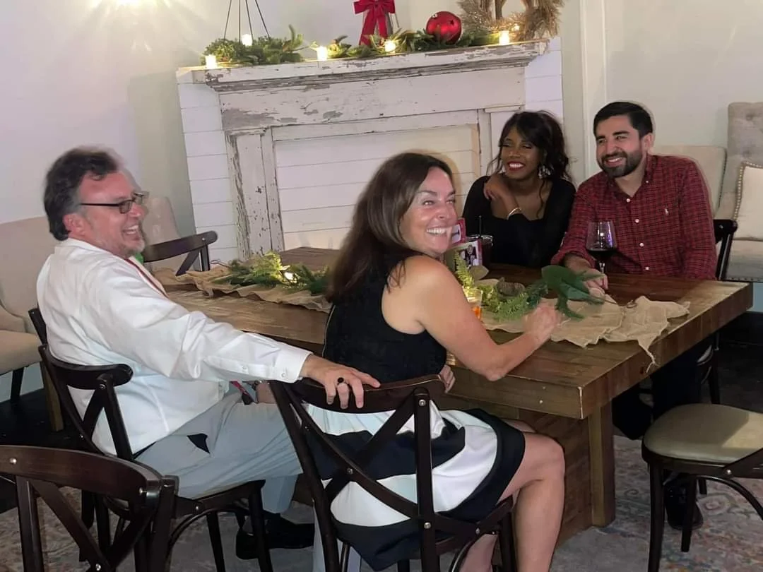 Four people sitting around a wooden table, smiling and laughing, with Christmas decorations on a mantelpiece behind them.