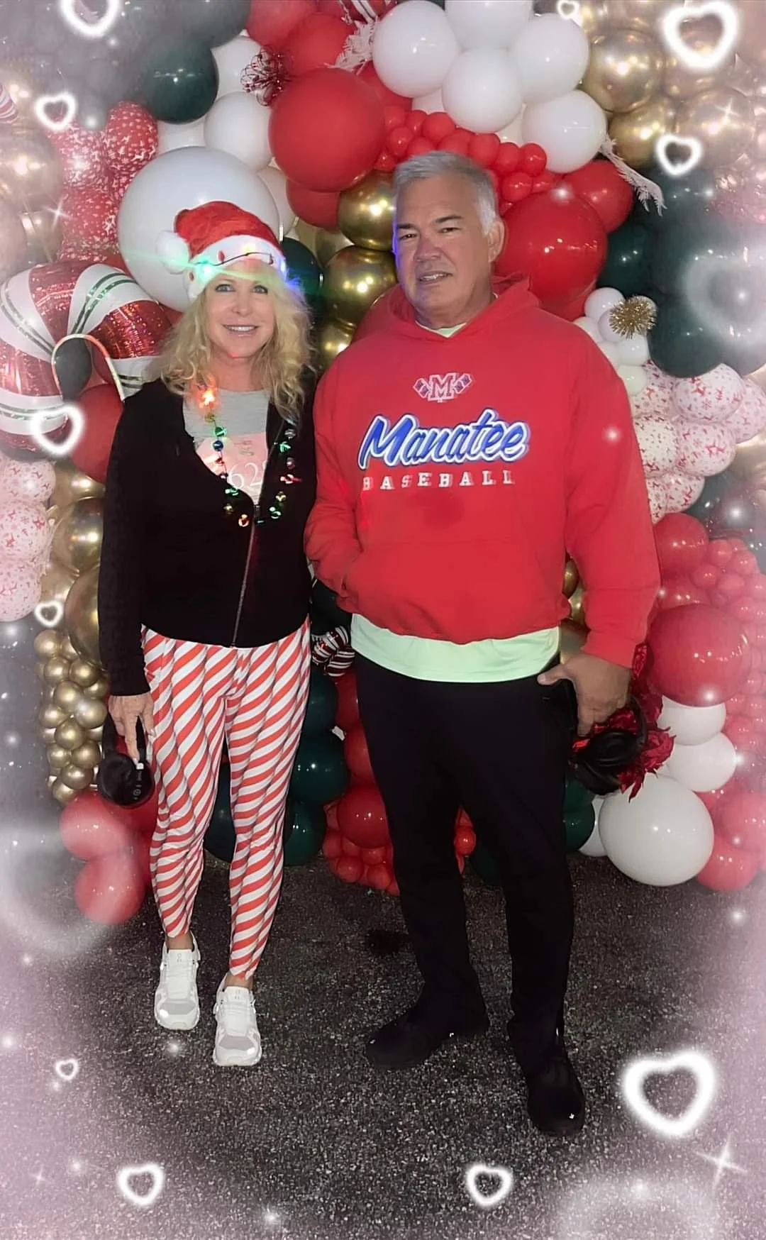 A woman and a man standing in front of a Christmas-themed balloon decoration. The woman is wearing a Santa hat, a black jacket, striped red and white pants, and white sneakers. The man is wearing a red hoodie with 'Manatee Baseball' written on it, an