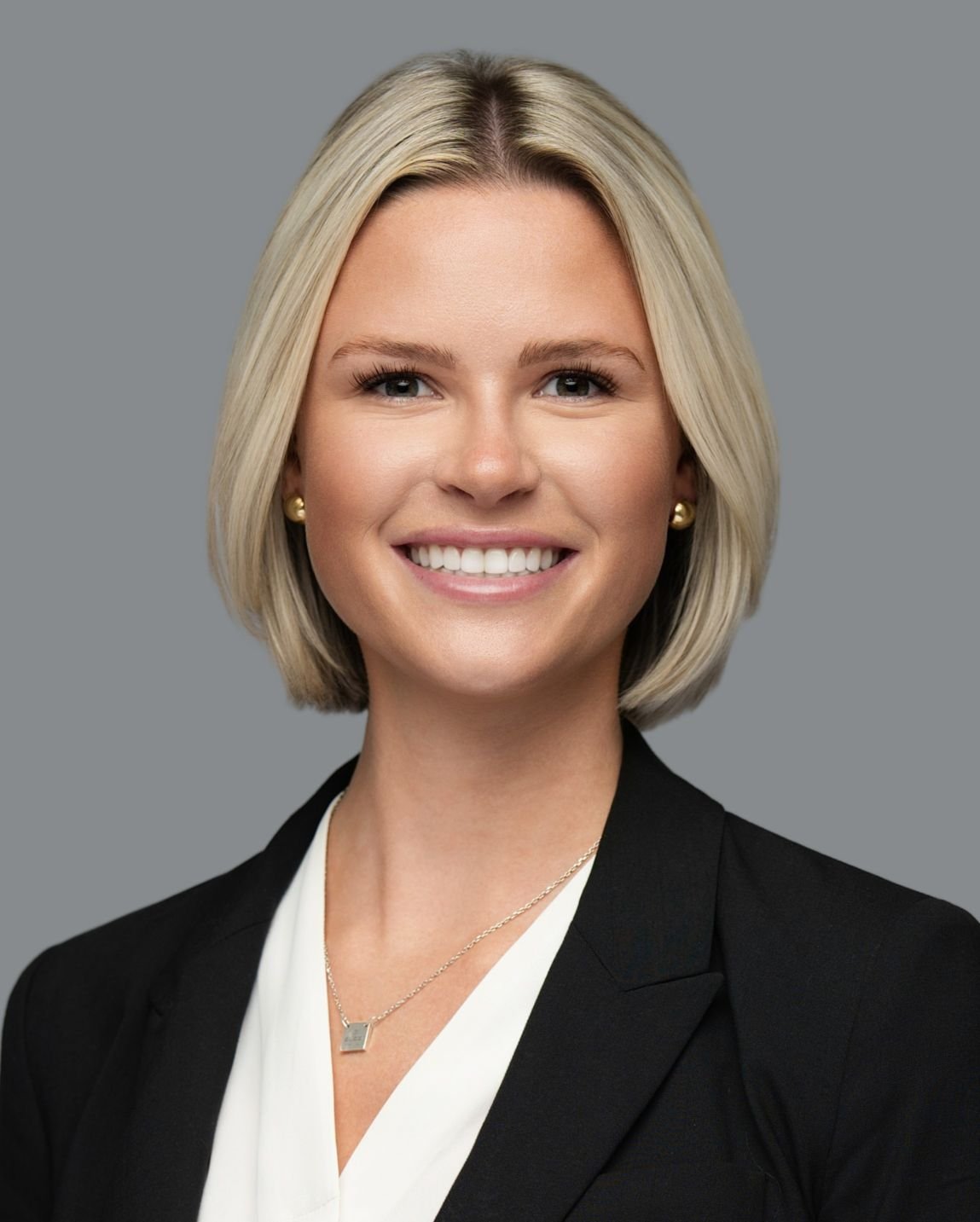 Portrait of a smiling blonde woman with shoulder-length hair, wearing a black blazer, white blouse, gold earrings, and a gold necklace, against a gray background.