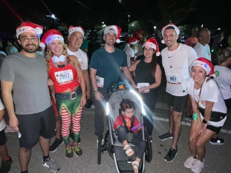Group of people at a nighttime Christmas-themed event, wearing Santa hats and festive attire, with a child in a stroller.