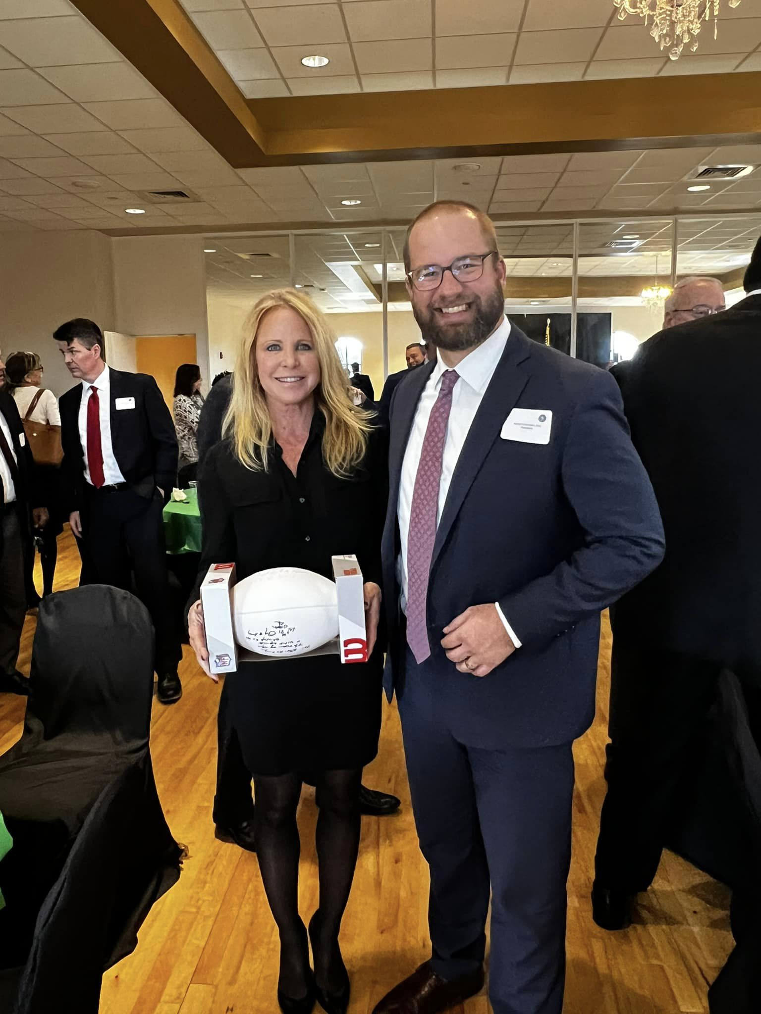 A woman with blond hair and a man with glasses and a beard smiling at a social event, holding a white box with writing on it. The woman is dressed in black and standing close to the man, who is dressed in a suit and tie. Other people are visible in t